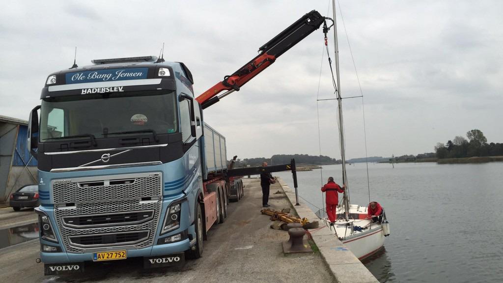 En lastbil fra Volvo med kran løfter en sejlbåd op fra vandet ved en kaj. En mand i rød dragt hjælper til.