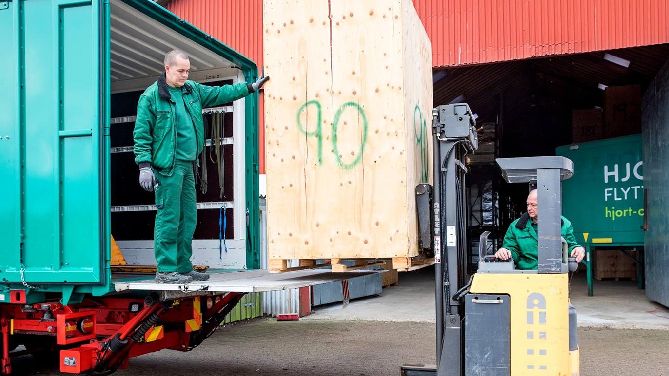 To mænd i grønne arbejdstøj, der arbejder med en gaffeltruck og en stor kasse i en lagerhal.