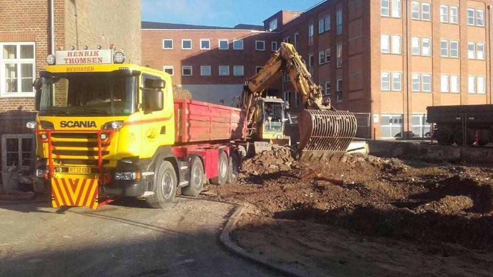 En gul Scania lastbil og en gravemaskine arbejder på en byggeplads ved en bygning.