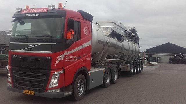 En rød Volvo lastbil med en sølvgrå tankvogn parkeret på en grusvej ved en bygning.