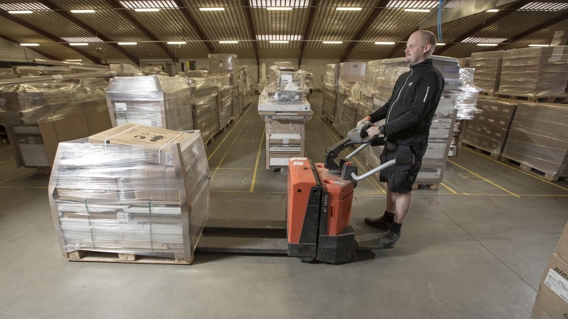En mand bruger en palleløfter til at flytte kasser i et lager fyldt med paller.