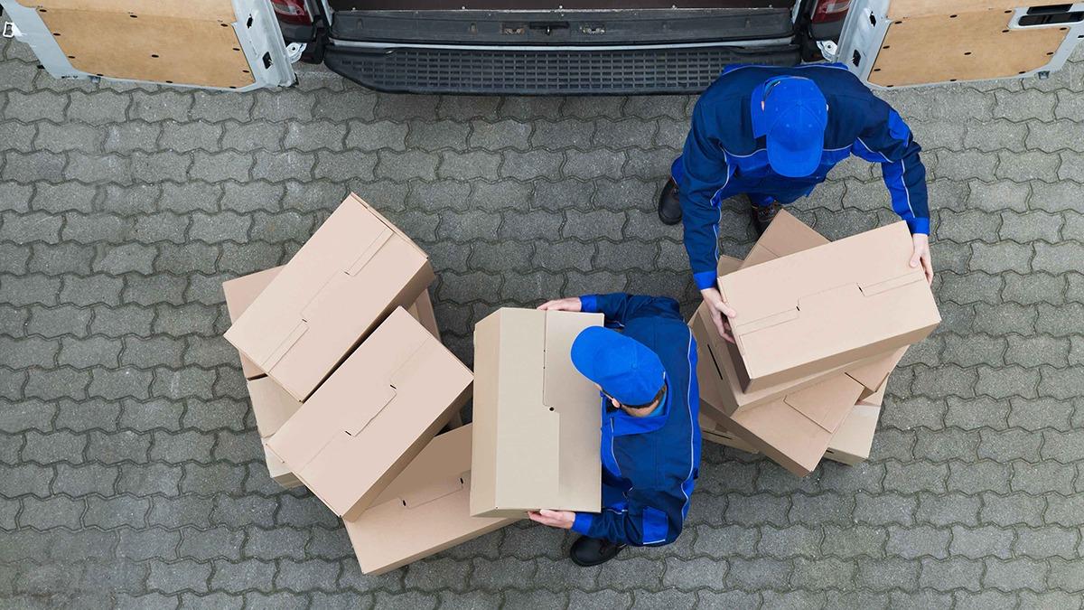 To personer i blå arbejdstøj bærer papkasser ved en varevogn på en brostenbelagt overflade.
