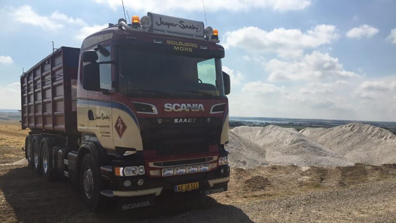 En Scania lastbil parkeret ved en grusgrav med en stor grushob i baggrunden under en blå himmel.