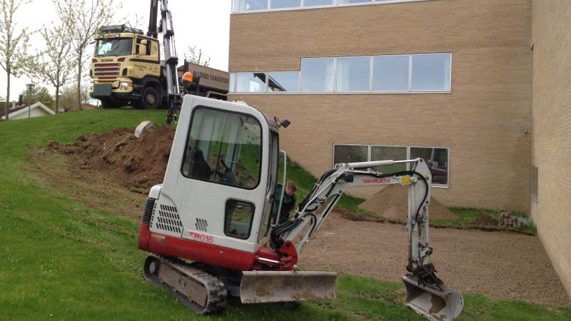 Gravemaskine arbejder på en skråning ved en bygning, mens en lastbil aflæsser materialer.