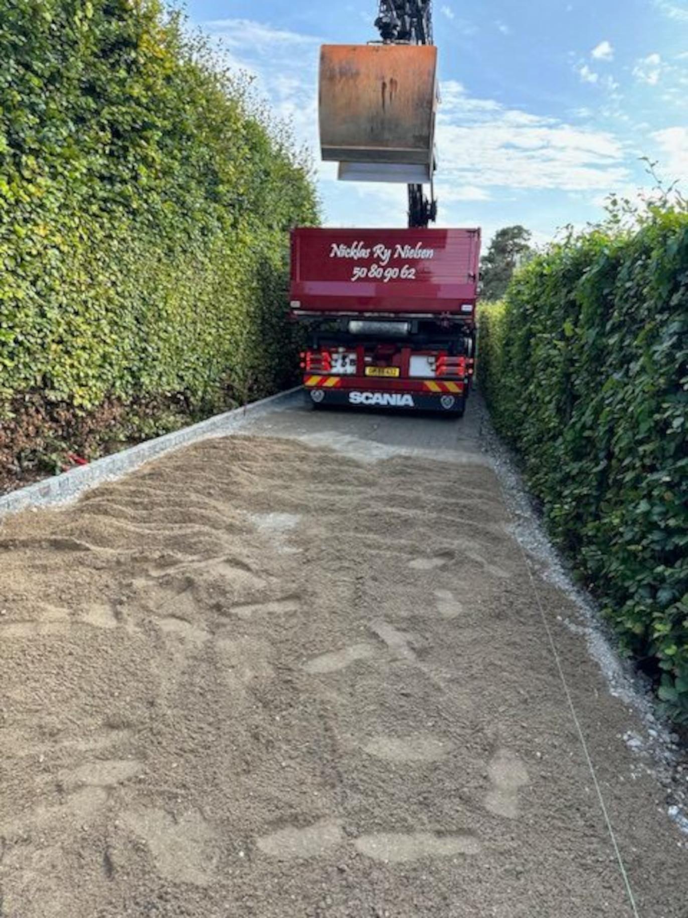 Lastbil med kran, der læsser sand på en grusvej omgivet af høje hække under en blå himmel.