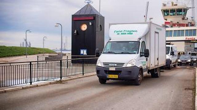 En hvid varevogn kører mod en færgeterminal med en stor klokke i baggrunden.