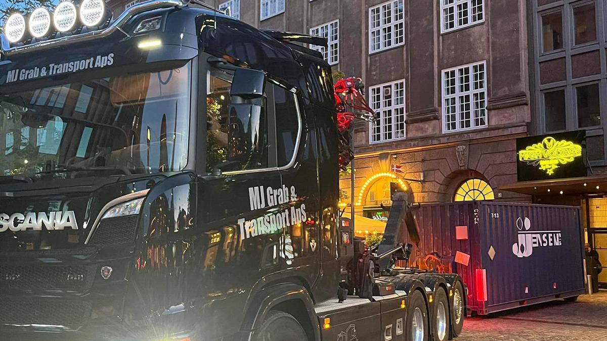 En sort Scania lastbil parkeret foran en bygning med lys og en container bagved.