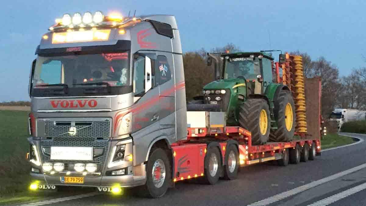 En Volvo lastbil med en stor traktor på en trailer kører langs en vej ved skumring.