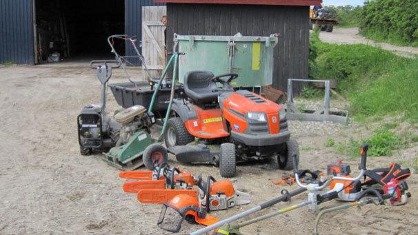 En samling af haveudstyr, herunder en plæneklipper, motorsave og en græstrimmer, ved en lade.
