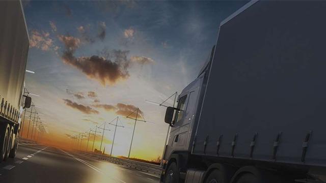 To lastbiler kører på en vej ved solnedgang med farverige skyer og elektriske master i baggrunden.
