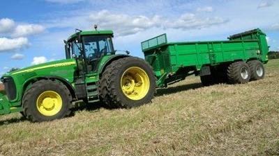 En grøn traktor trækker en stor vogn på en mark under en blå himmel med skyer.