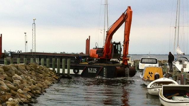 En gravemaskine arbejder ved havnen, mens både ligger fortøjet langs kajen.
