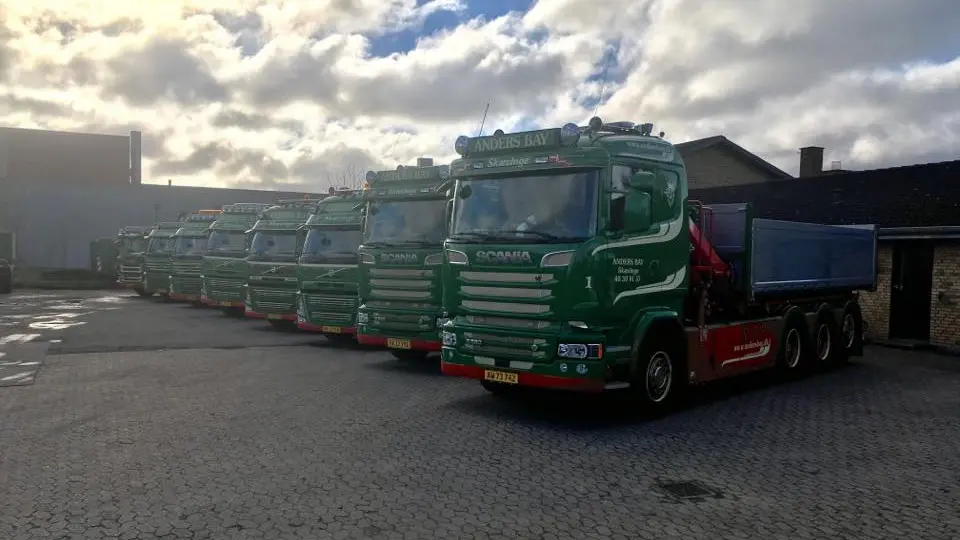 Flere grønne Scania lastbiler parkeret i en gård under en skyet himmel.