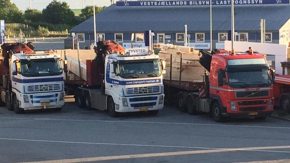 Tre lastbiler parkeret på en havn med trælast på ladet og en bygning i baggrunden.