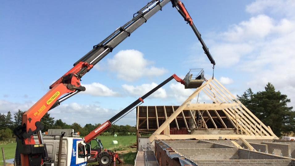 Kraner løfter tagkonstruktion på et hus under byggeri med blå himmel og grønne marker i baggrunden.