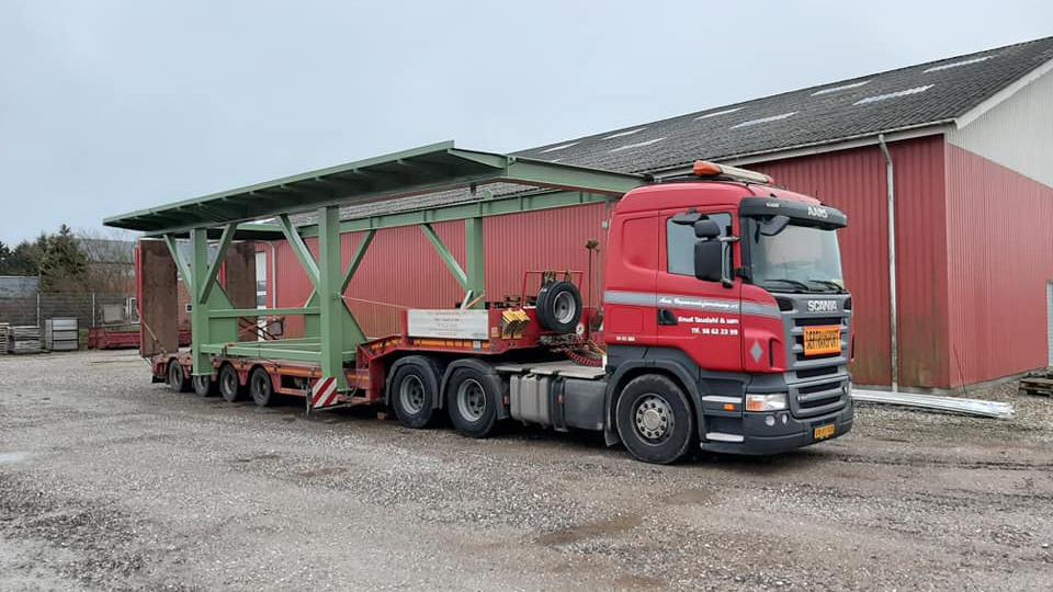 En rød lastbil med en stor grøn trailer parkeret ved en bygning i en gård.