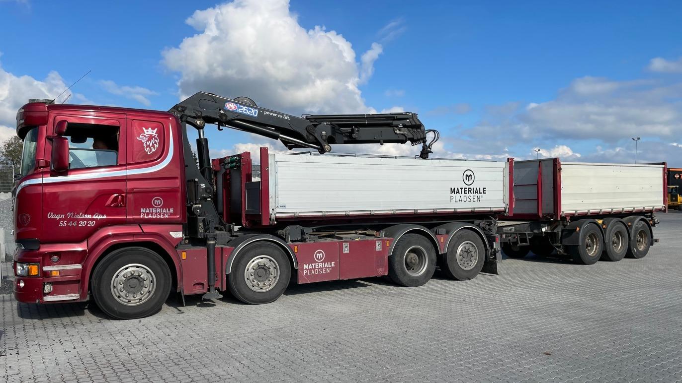 En rød lastbil med kran, der transporterer to containere, parkeret på en brostenbelagt plads.