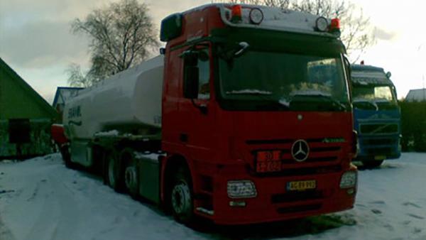 En rød tankvogn parkeret i sneen med en hvid tank og træhus i baggrunden.