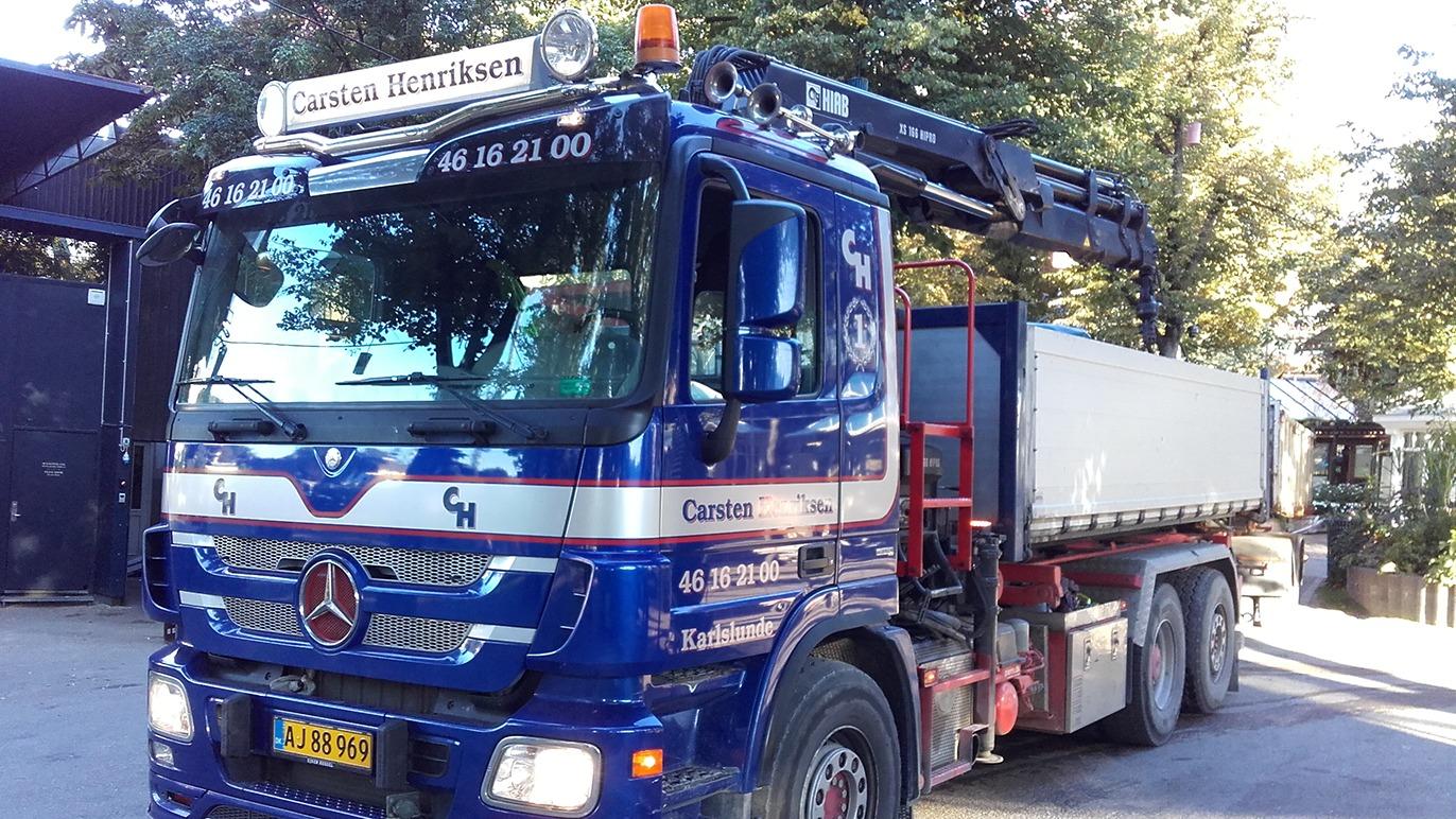 En blå lastbil fra Mercedes med kran og trailer, parkeret i et skovområde.