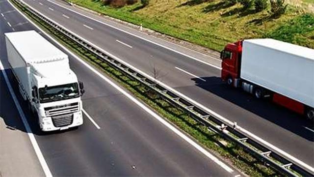 To store lastbil og en rød lastbil kører på en motorvej med græs og træer i baggrunden.