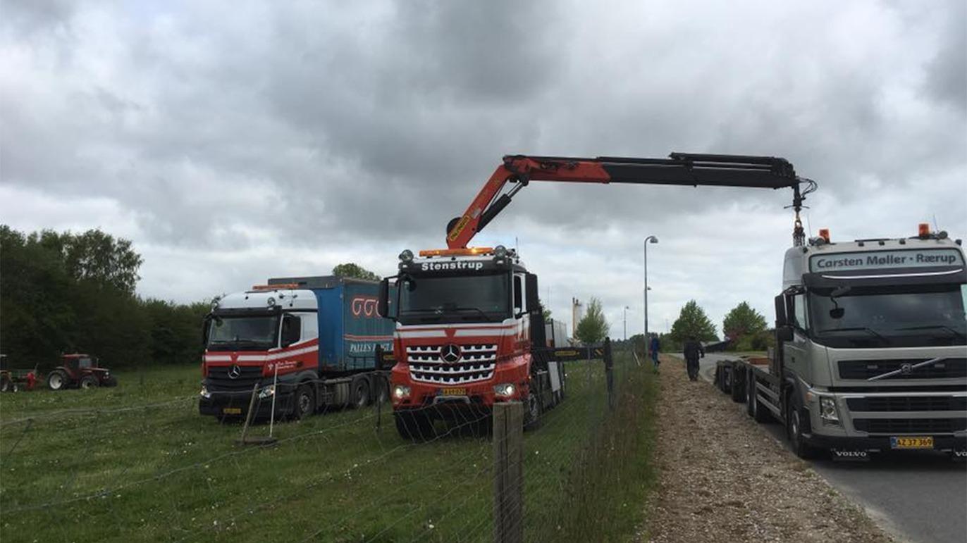 To lastbil med kran arbejder på en markvej, mens en anden lastbil holder parkeret i baggrunden.