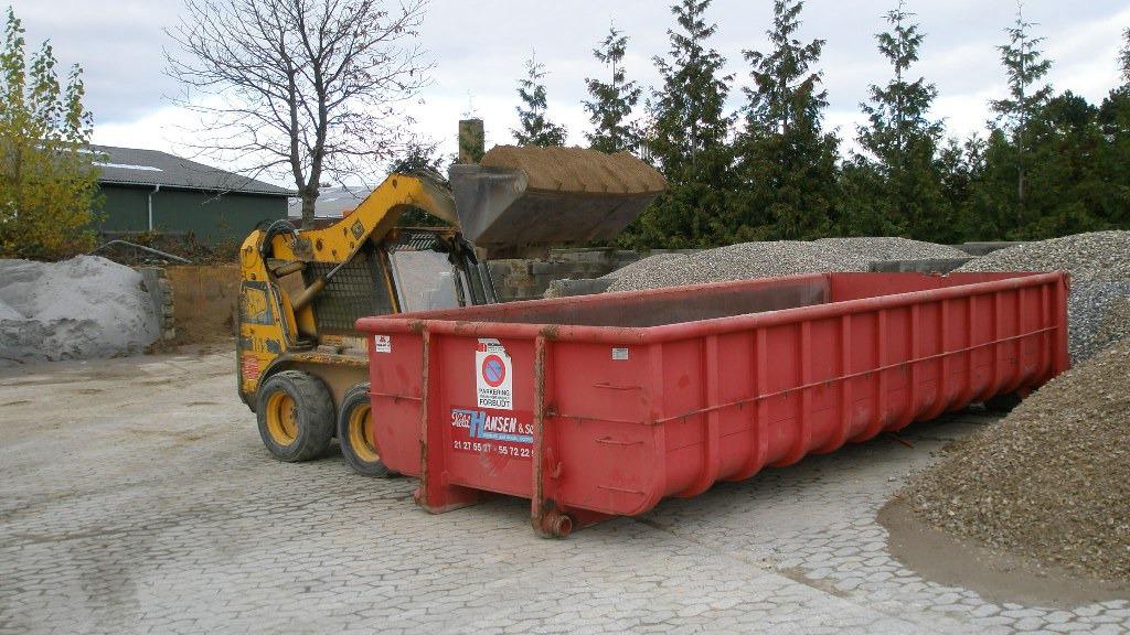 En gult gravemaskine læsser materialer i en rød affaldscontainer på en byggeplads.