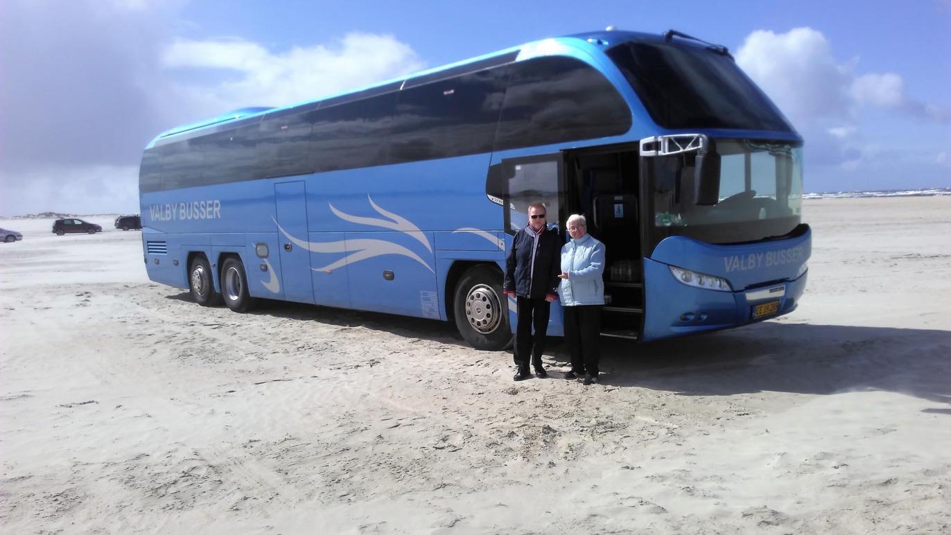 En blå turistbus parkeret på stranden med to personer stående foran.