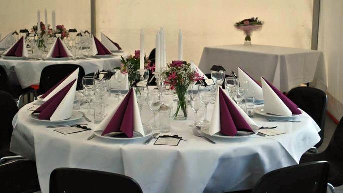 Elegant festbord med hvide duge, lilla servietter, glas og blomsterdekorationer.