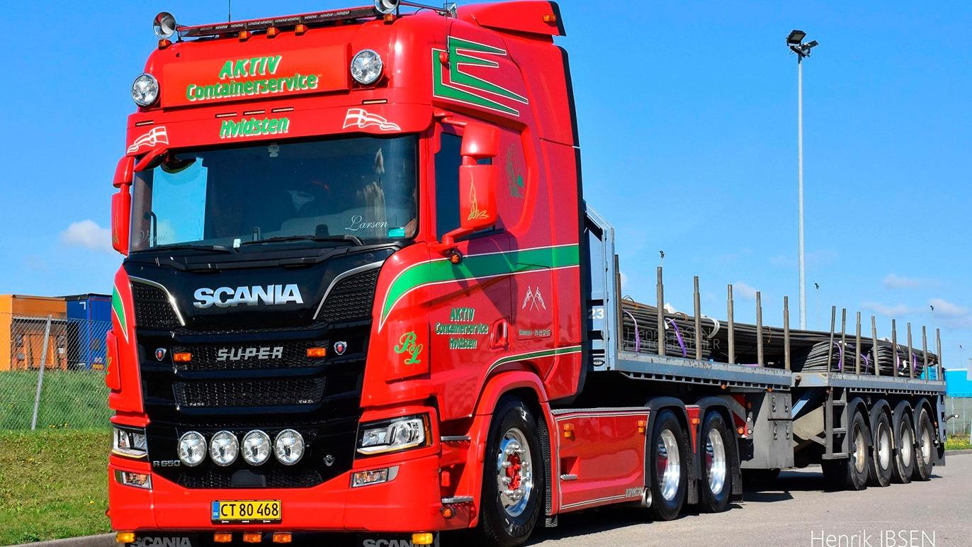 En rød Scania lastbil med en flad trailer parkeret i et industriområde under en blå himmel.