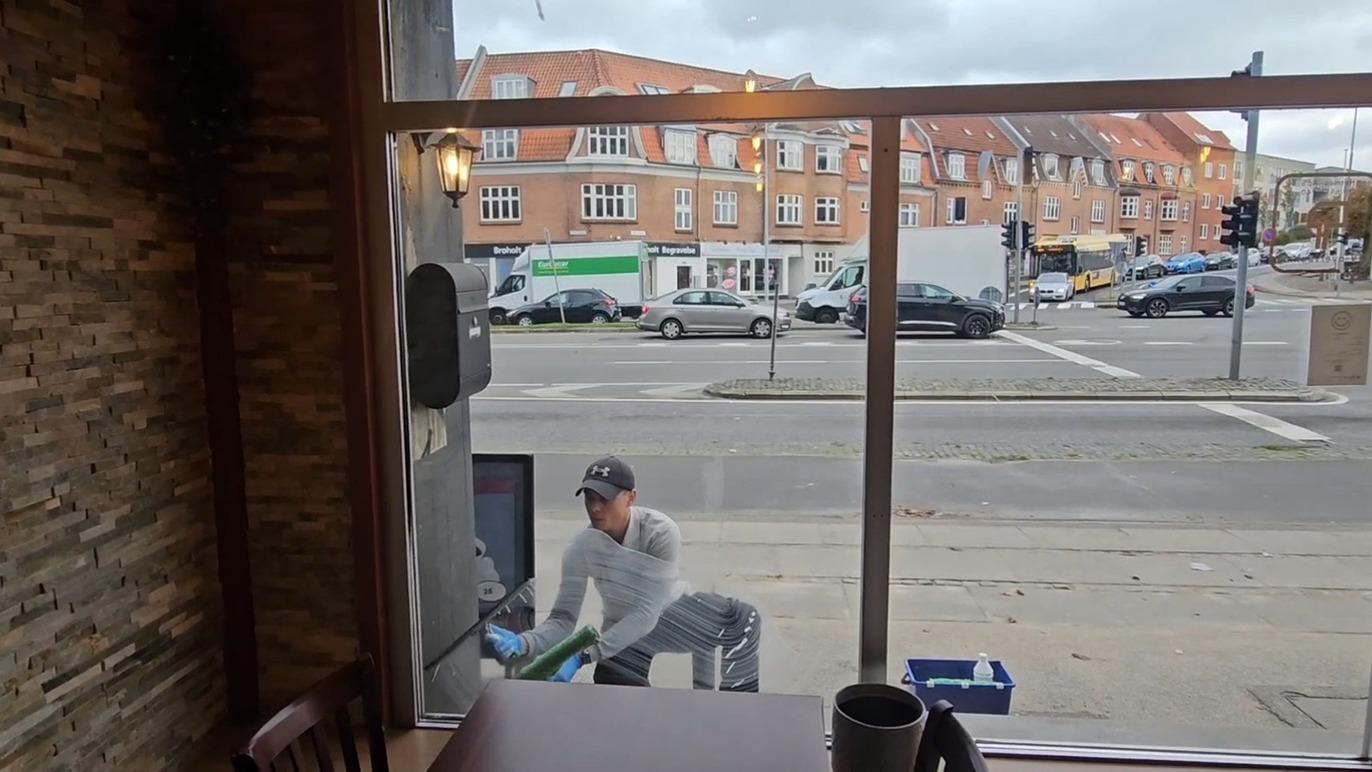 En mand står udenfor en café og rengør vinduet, mens biler kører forbi på gaden.