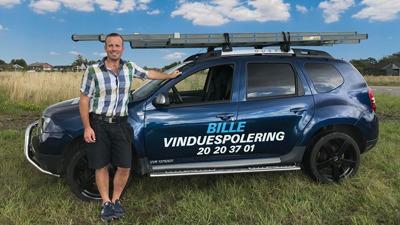 En mand står ved siden af en blå bil med teksten "BILLE VINDUESPOLERING" på siden, i et åbent landskab.