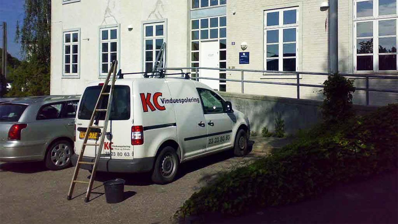 En hvid varevogn med teksten "KC Kenderespartiet" parkeret ved en bygning med vinduer og en stige.