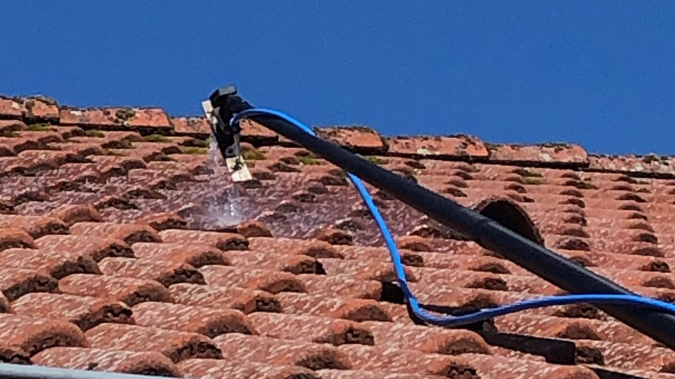 Vandstråle fra en højtryksrenser, der rengør et tag med røde teglsten under en blå himmel.