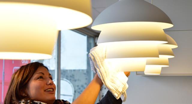 En kvinde rengør en moderne lampe i en lys butik med flere lamper i baggrunden.