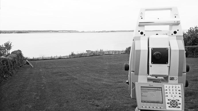 Et totalstation står på en græsplæne med udsigt over en sø og en grå himmel i baggrunden.
