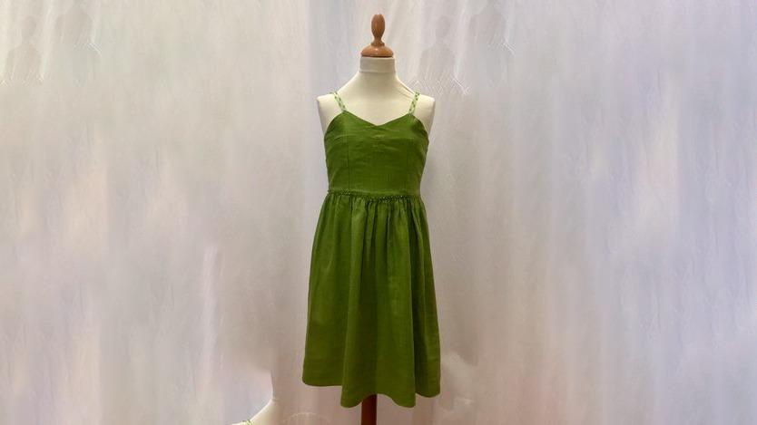 Grøn sommerkjole med spaghettistropper hængende på en mannequin foran en lys baggrund.
