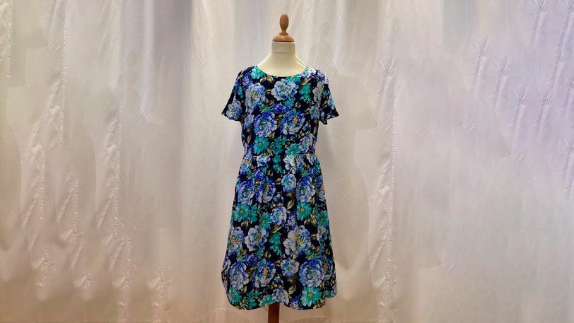 En blomsterkjole med blå og grønne nuancer hængende på en mannequin foran en lys baggrund.