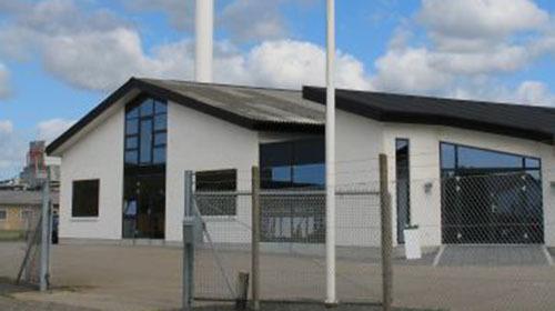 Moderne bygning med store vinduer og sort tag, omgivet af hegn og med blå himmel i baggrunden.