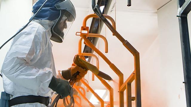 En person i beskyttelsesdragt sprøjter orange maling på metalrammer i et malerværksted.