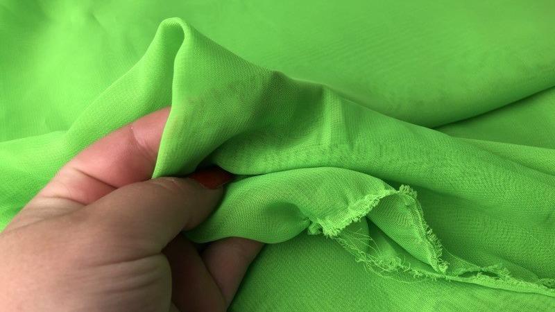 Hånd, der holder et stykke lys grøn chiffonstof med bløde folder og let struktur.