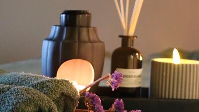 Et afslappende spa-setup med håndklæder, duftlys, en vase med lavendel og en sort beholder.