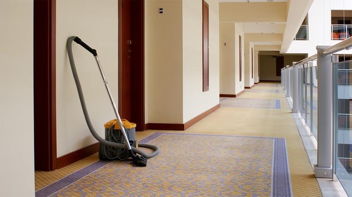 En støvsuger står på et hotelgang med beige vægge og et mønstret tæppe.