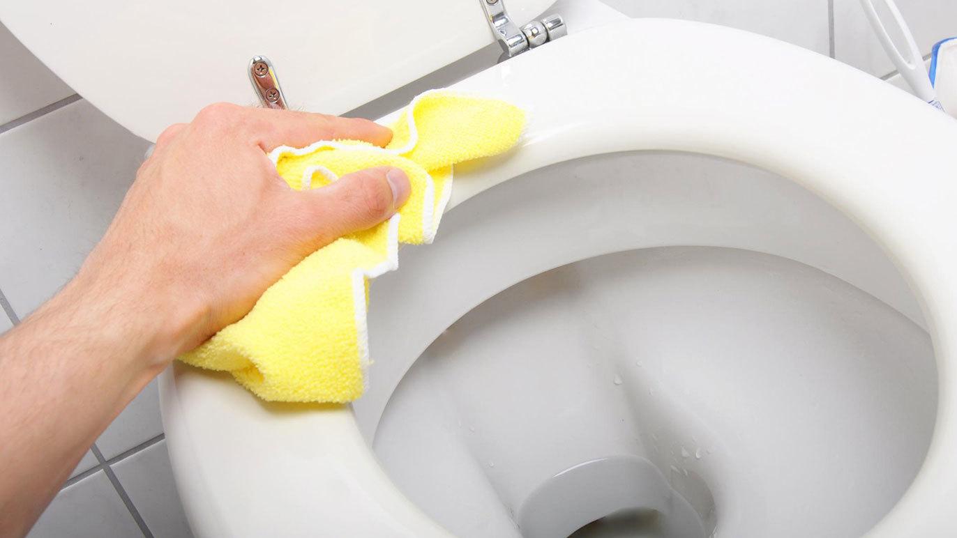 En hånd, der rengør en toiletbræt med en gul klud i et badeværelse.