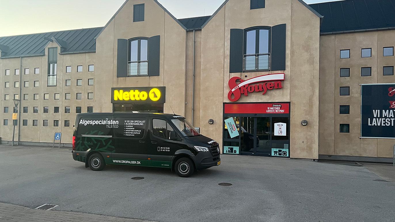 En sort varevogn parkeret foran Netto og en butik med skiltet "Spar" i en bygning.