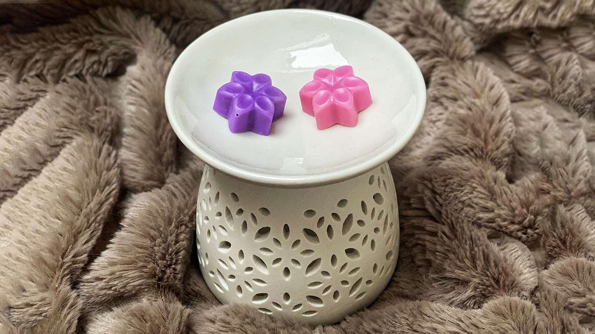 En hvid duftlampe med to farverige voksblomster, en lilla og en pink, placeret på en blød, beige overflade.