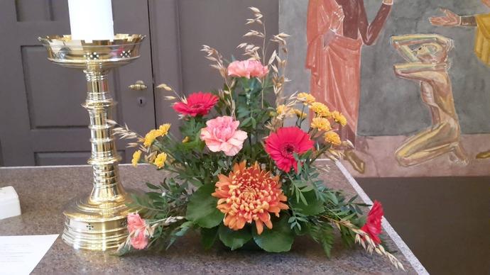 En smuk blomsterdekoration med farverige blomster som gerbera, nelliker og chrysanthemum ved en lysestage.