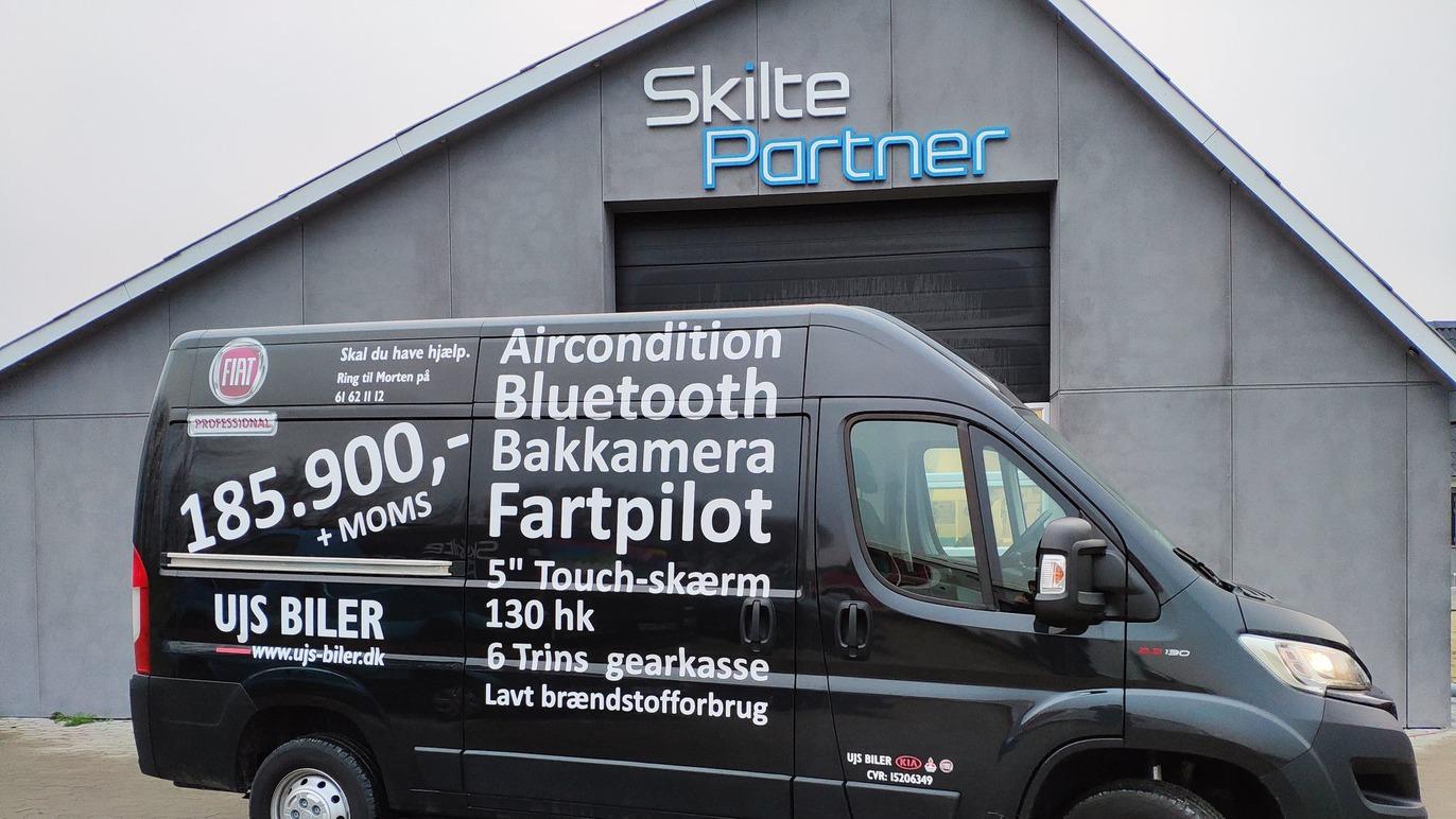 En sort varevogn med tekst om funktioner som aircondition og bakkamera, parkeret foran en bygning med "Skilte Partner".
