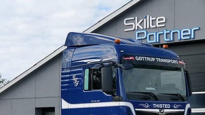 Blå lastbil fra Gottrup Transport parkeret foran Skilte Partner bygning med skyet himmel.