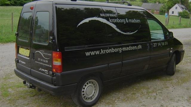 Sort varevogn med logo og hjemmeside på siden, parkeret på en grusvej i et grønt landskab.