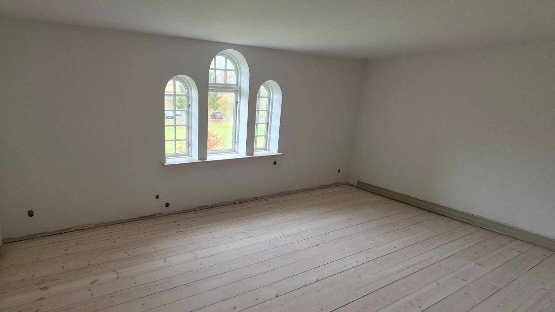 Lyst og rummeligt værelse med hvid vægfarve og store vinduer, der giver naturligt lys.
